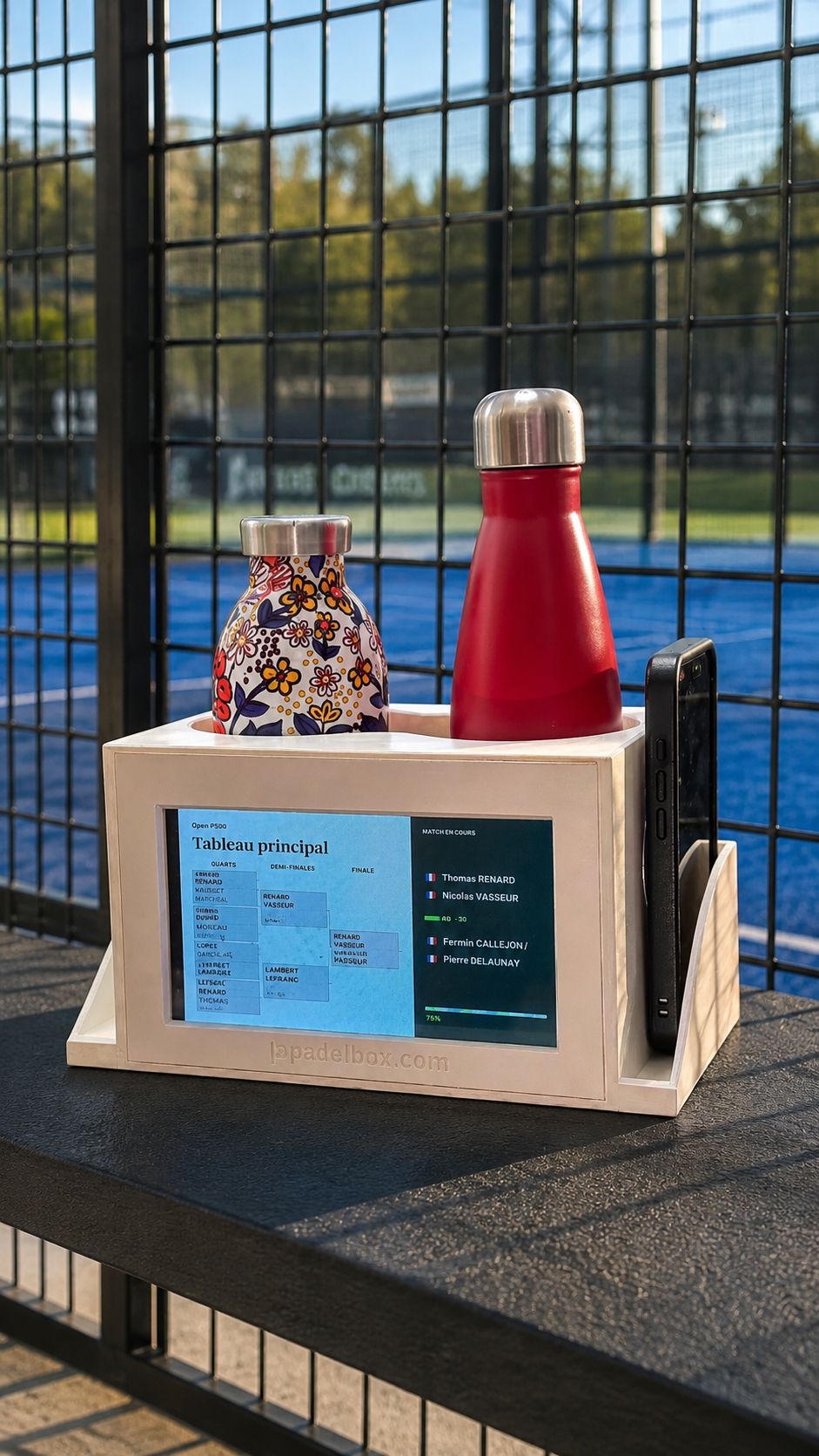 La PadelBox installée au grillage d'un terrain : gourdes et téléphone rangés, l'écran affiche le tableau du tournoi et le match en cours.